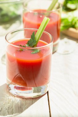 Two glasses with tomato juice, celery and parsley stand on a wooden table in white. Stalks of celery sliced on a cutting board in the background.