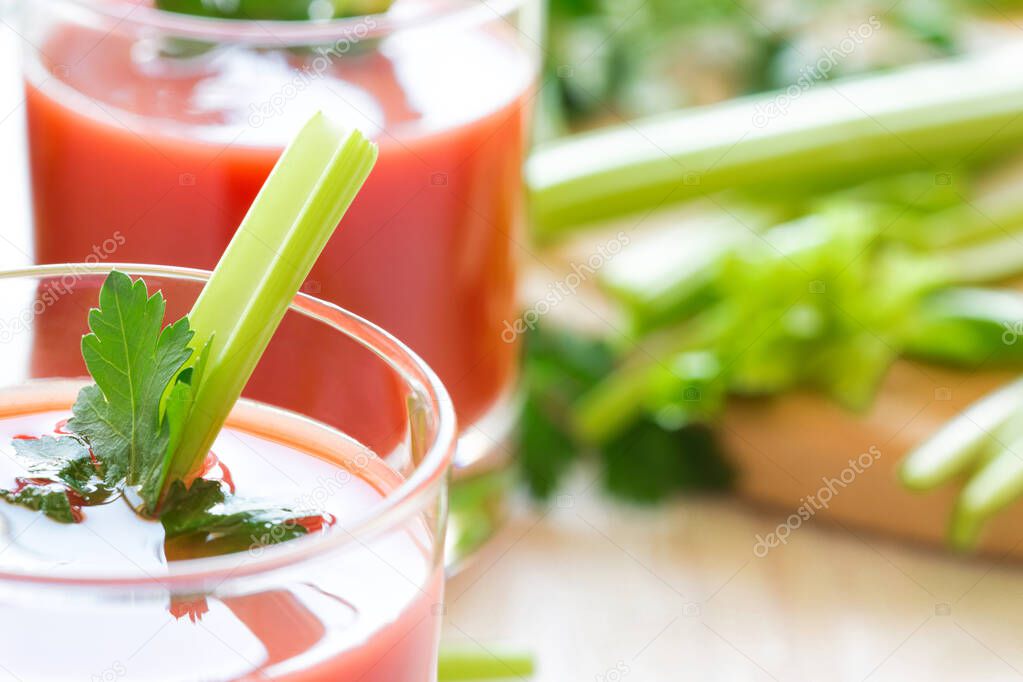 Two glasses with tomato juice, celery and parsley stand on a wooden table in white. Stalks of celery sliced on a cutting board in the background.