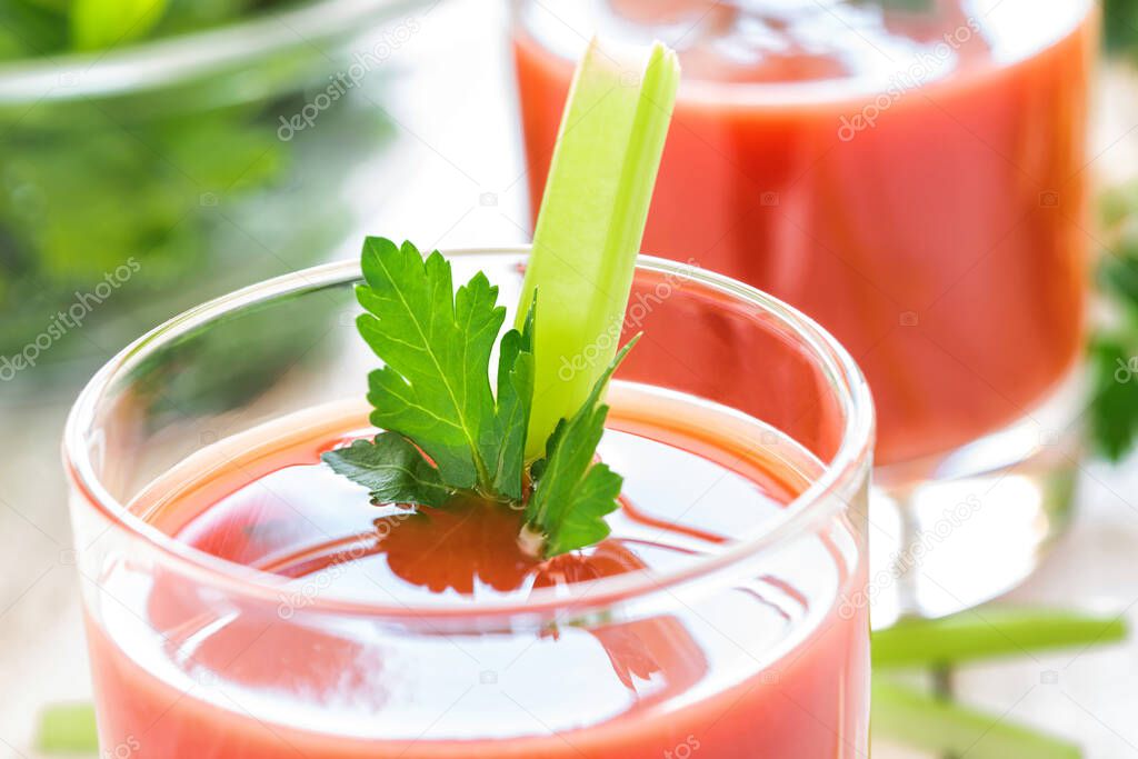 Two glasses with tomato juice, celery and parsley stand on a wooden table in white. Stalks of celery sliced on a cutting board in the background.
