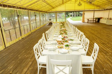 Ahşap zeminli ve beyaz sandalyeli geniş çadırda kurumsal etkinlik için hazırlanmış zarif uzun ziyafet masası..