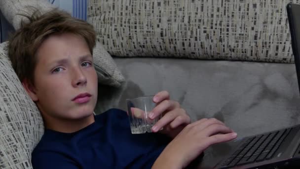 Мальчик со стаканом воды и ноутбуком смотрит в камеру. — стоковое видео