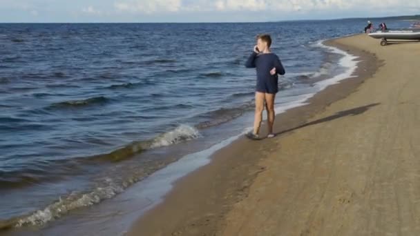 เด็กชายผิวขาวเดินไปตามชายหาดและพูดจากสมาร์ทโฟน . — วีดีโอสต็อก