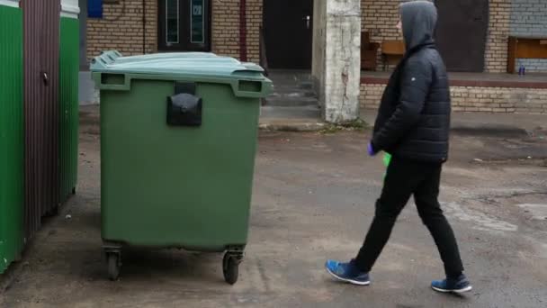 Un chico europeo de 15 años arroja una bolsa de plástico en una bolsa de plástico verde a un cubo de basura. — Vídeo de stock