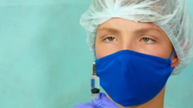 Maskeli genç bir beyaz erkek doktor eldivenli eliyle bir şırınga tutuyor..