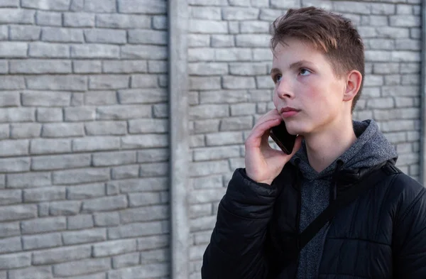 En tonårspojke med mörkt hår i svart jacka som står på gatan med en mobiltelefon i händerna. — Stockfoto