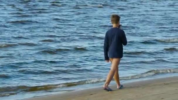 Un chico con una camiseta azul camina por la orilla del mar. — Vídeo de stock