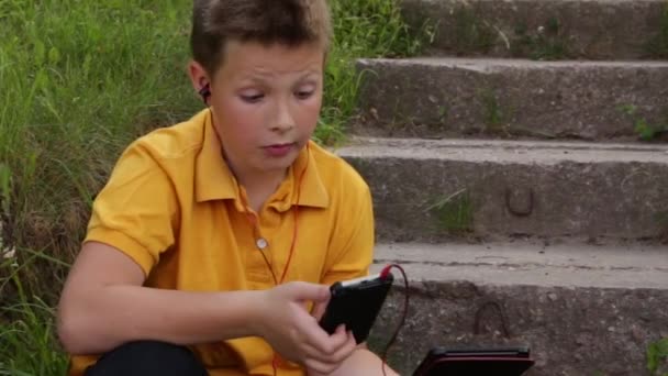 Ein Junge sitzt auf den Stufen und spielt mit zwei Geräten. — Stockvideo