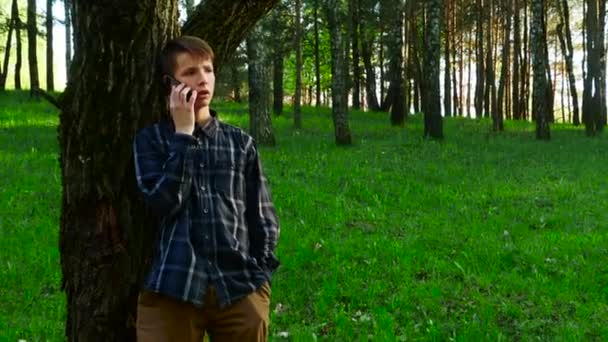 Un niño de 13-17 años se para en el parque cerca de un árbol y habla por teléfono. — Vídeos de Stock