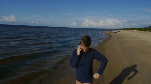 Um menino falando ao telefone está andando ao longo da praia do mar. — Vídeo de Stock