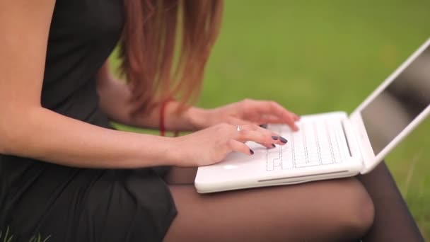 Jovem mulher digitando em um laptop sentado na grama verde — Vídeo de Stock