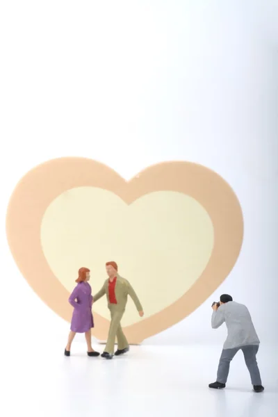 Miniatura de um fotógrafo tirando fotos de um jovem casal com um coração rosa e amarelo como fundo — Fotografia de Stock