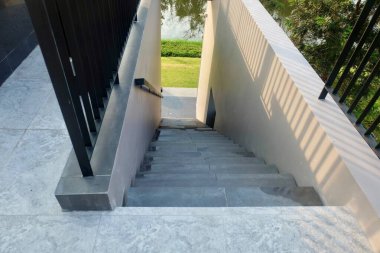 Yüksek beton duvarlar arasından inen minimalist koyu gri merdiven görüntüsü. Adımlar yeşil çimlere çıkar, güçlü gölgelerle vurgulanır..