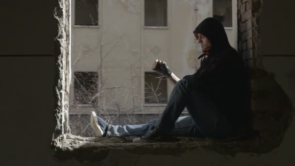 Jeune homme déprimé à capuchon assis sur le cadre de fenêtre d'un bâtiment abandonné — Video