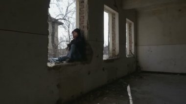 Terk edilmiş bir binada pencere çerçevesi üzerinde oturan kukuletalı depresif genç adam