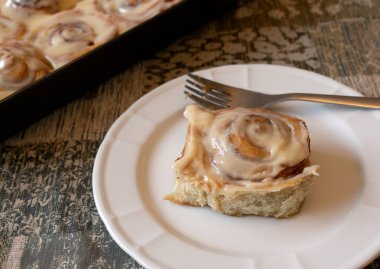 Krem peynirli tarçın ruloları, arka planda çatal ve tavayla beyaz tabağın üzerinde. 