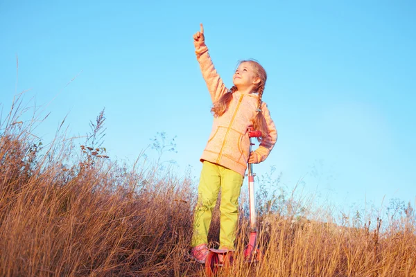 Liten flicka i ljusa kläder med skoter visar ett finger till himlen — Stockfoto