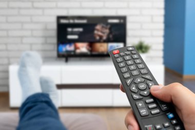 Televizyon izleyen bir adam, kanepeye uzanmış, bacakları masanın üstünde. Oturma odasında uzaktan kumandalı biri var.