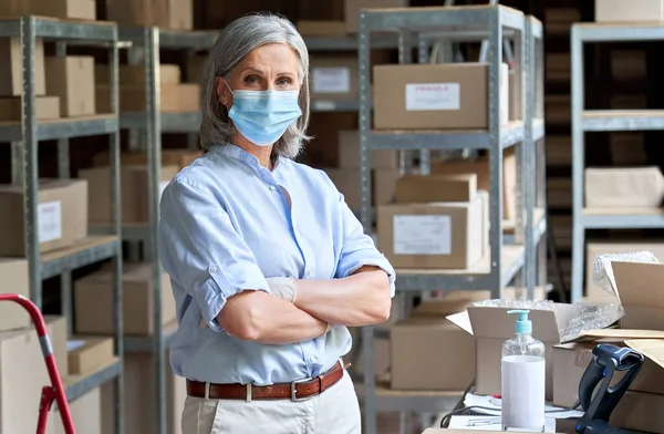 Mature woman entrepreneur wearing face mask standing in warehouse, portrait.