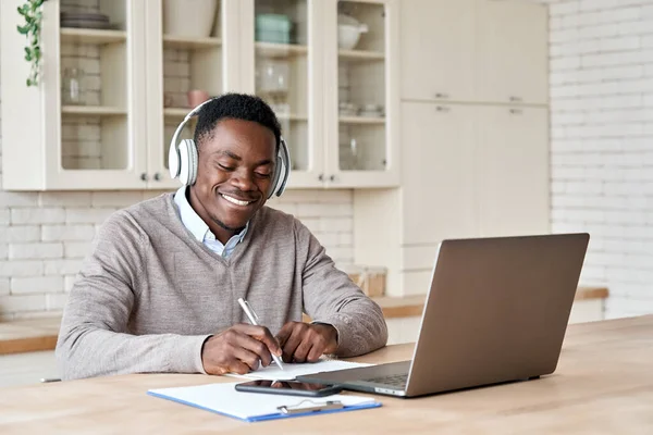 Black male student wearing headphones elearning on laptop at home office.