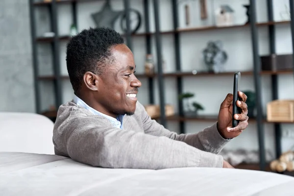 Happy african young man using mobile apps sitting on couch at home.