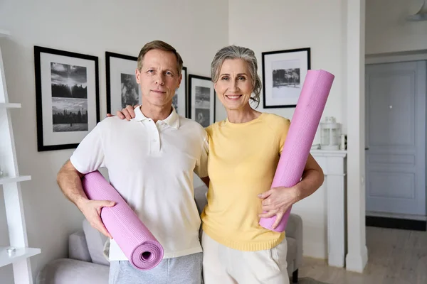 Happy fit old couple hugging, looking at camera, holding yoga mats at home.
