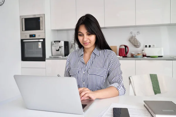 Indian woman working online from home office sitting at table using laptop.