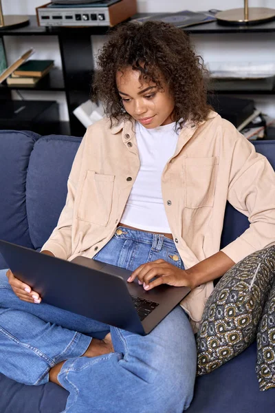 African American female student sitting on soft sofa at home using pc laptop.