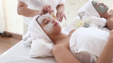 Güzellik uzmanları genç Asyalı kadınların yüzüne beyaz yüz maskesi takmak için spatula kullanırlar. Kamerayı yakınlaştırarak.