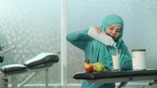 緑のヒジャーブの若い女性ムスリムたちがフィットネスルームのベンチで牛乳をグラスに注いでいます スポーツ ボディービル フィットネス 人々のコンセプト — ストック動画
