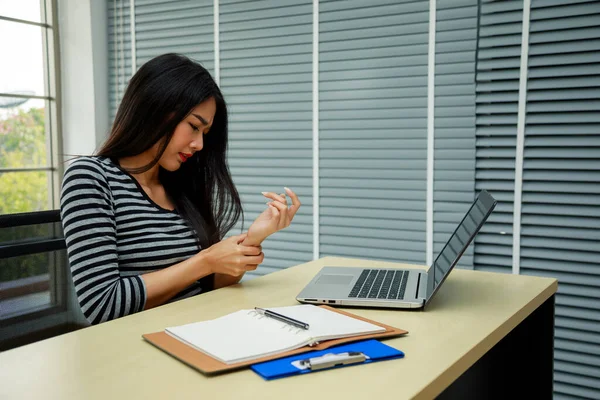 Genç Asyalı Kadın odada bilgisayarla bileğini tutarak acı çekiyor. Ofis Sendromu el ağrısı hastalığı
