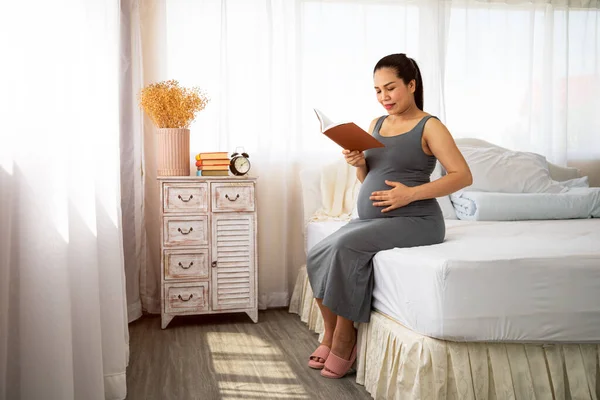 Happy Asia pregnant woman reading book and sitting on white bed. Relaxation give birth mother