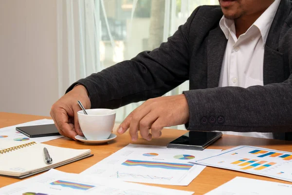 Asian young businessman holding move coffee cup checking sales data chart sheet on desk in office