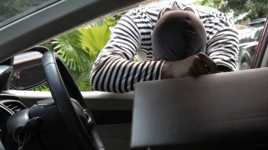 Açık pencereden içeri uzanan çizgili tişörtlü maskeli bir adam. Aracın derinliklerine doğru eğiliyor, muhtemelen bir şey çalmak için.