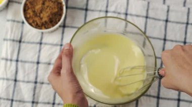 Yukardan yukarı yakın plan, cam kasede hamuru çırpıyor, kareli kumaş üzerinde, basit pişirme malzemeleriyle çevrili, ev yapımı tatlı hazırlama anı