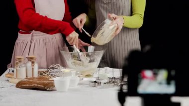 İki kadın birlikte yemek pişirirken cam un kasesine kurabiye hamuru kazırlar. Mutfak masasında pişirme malzemeleri, mutfak gereçleri ve kurabiye keskisi var.