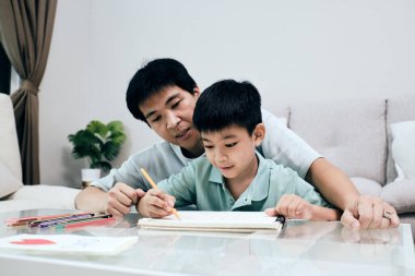 Asyalı bir baba, evde çizim yaparken çocuğuna rehberlik ediyor, yaratıcılığı, öğrenme gelişimini ve sıcak ve besleyici bir ortamda pozitif aile etkileşimini vurguluyor.