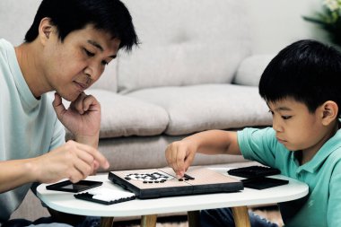 Asyalı baba ve oğul, küçük beyaz masada stratejik siyah-beyaz masa oyunu oynarken derinden odaklandılar; aile bağları, eleştirel düşünme ve eğitim.