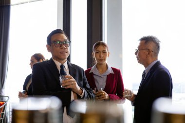 İş dünyasından profesyoneller, iş birliği iletişimini ve profesyonel etkileşimi gösteren pencerelerin yakınındaki kurumsal ağ etkinliğinde içkilerle birlikte duruyorlar.