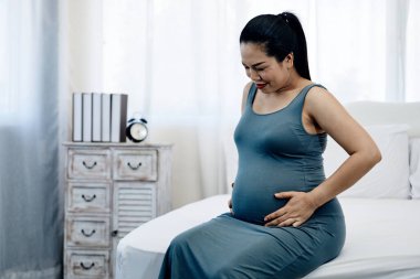 Kolsuz mavi elbiseli hamile bir Asyalı kadın elleri karnına hafifçe otururken gülümsüyor ve aşağı bakıyor sıcak, bağlantı ve annelik sevincini iletiyor.