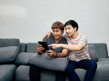 İki Asyalı adam modern gri koltukta oturuyor, tablet kullanırken gülümsüyor ve etkileşimde bulunuyor, biri çizgili gömlekli, ekranı işaret ediyor ve diğer gri gömlekli adamı işaret ediyor.