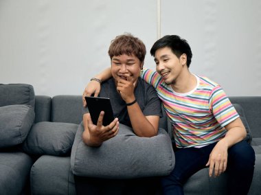 İki LGBTQ Asyalı adam gri koltukta oturmuş tablete bakarken sıcacık gülümsüyor, biri çizgili gömlekli, diğeri gri gömlekli.