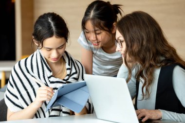 Genç öğrenci, öğretmenleri ve kızlarıyla birlikte tablet kullanıyor. Sınıfta dizüstü bilgisayarda birlikte okuyorlar. Çocuklara dijital eğitim konseptinde rehberlik eden yetişkin bir öğretmen.