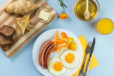 Nefis bir kahvaltı. Tereyağı, portakal suyu, reçel, bal, kruvasan, bıçak, çatal ile servis edilir. Sarı, mavi. Mavi arkaplan.