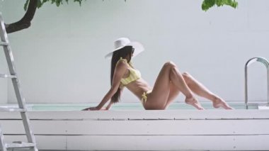 Güneş gözlüklü, beyaz şapkalı, şapkalı ve havuz kenarında güneşlenen genç bir kadın..
