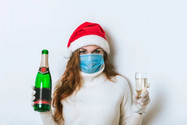 Christmas portrait of woman with medical protective facial face mask in studio with isolated background. Girl wear red Santa hat is holding drink champagne and glass. New year concept.