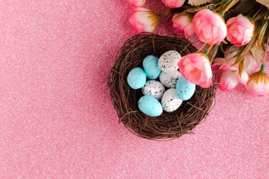 Yuvada renkli bıldırcın yumurtaları ve pembe parlak arka planda bir buket çiçek. Paskalya tatili konsepti
