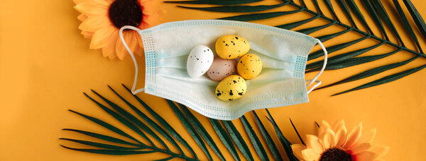Quail eggs in face medical mask and palm branches on yellow background. Easter holiday during the coronavirus pandemic