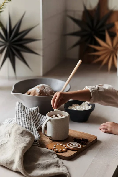 Küçük bir kızın eli bir fincan kahveye marşmelov koyar. Pencerenin yanında bir masa. Çok atmosferik ve rahat bir Noel fotoğrafı.