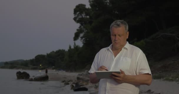 Старший чоловік використовує планшетний ПК на пляжі в кінці вечора — стокове відео