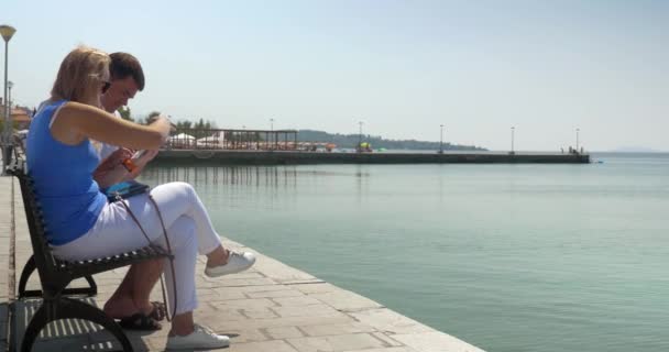 Hombre y mujer usando almohadilla sentados en banco frente al mar — Vídeos de Stock